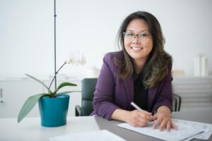 Asian woman with glasses smiles while working at a desk with documents and a potted plant.